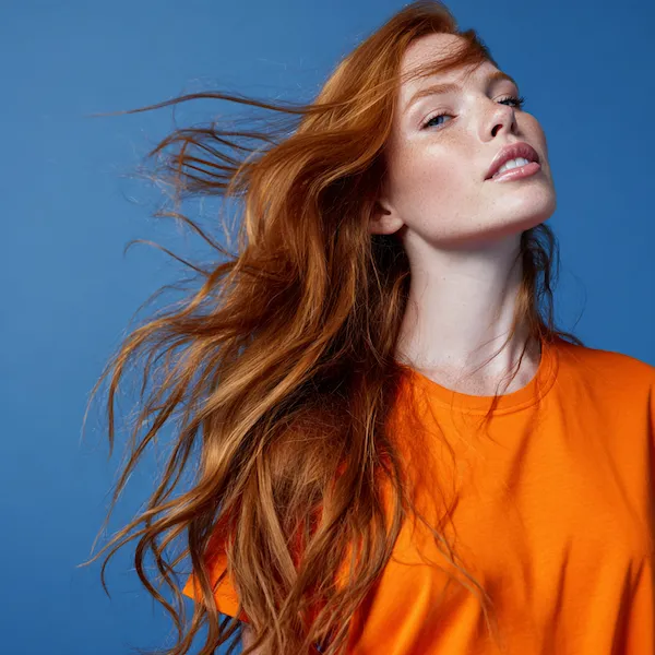 Junge Frau mit langen, natürlichen kupferroten Haaren in Bewegung vor blauem Hintergrund – Ergebnis professioneller Haarverlängerung