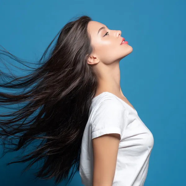 Junge Frau mit langen, glänzenden dunkelbraunen Haaren in Bewegung vor blauem Hintergrund – natürliche Haarverlängerung