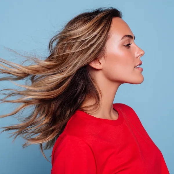 Junge Frau mit langen, braunen Haaren und blonden Highlights in Bewegung vor hellblauem Hintergrund – Inspiration für Balayage und Strähnenfarben